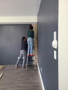 two people standing on ladders painting the walls in a room with wood flooring