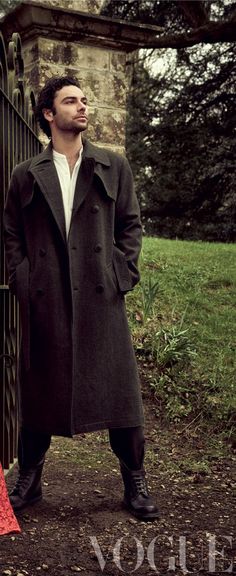 a man standing next to a gate wearing a long coat and red dress pants with his hands in his pockets