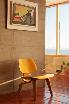 a yellow chair sitting on top of a red tile floor next to a painting and potted plant