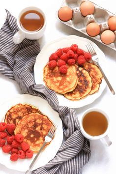 pancakes with raspberries and syrup on white plates next to cups of tea, eggs and spoons