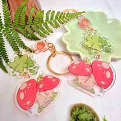 three keychains with different designs on them sitting next to a fern leaf and potted plant