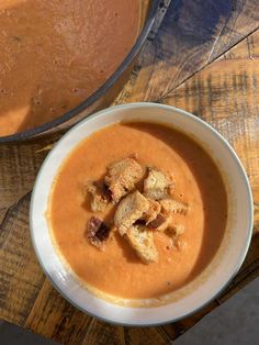 a bowl of soup with croutons in it on a wooden table next to a pot of soup