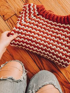 someone is holding up a red and white crocheted bag on the floor next to their feet