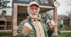 an older man with white hair and beard standing in front of a house holding his fist up