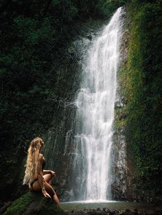 a woman sitting on top of a mossy rock next to a waterfall in the forest