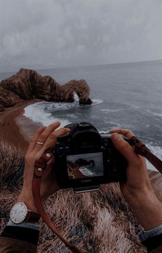 a person holding up a camera to take a photo near the ocean with cliffs in the background