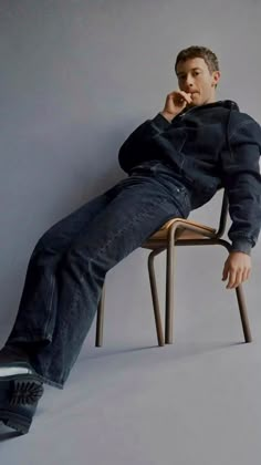 a man sitting on top of a wooden chair next to a white wall with his hand under his chin