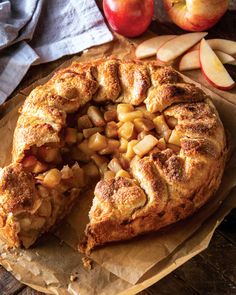 an apple pie with one slice cut out and some apples around it on a piece of wax paper