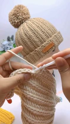 two hands are holding yarn in front of a doll's head with a crochet hat on it