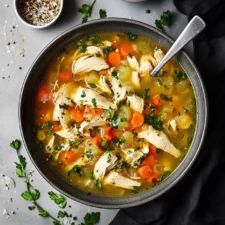 a bowl of chicken and vegetable soup on a table with two bowls of seasoning