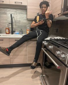 a man sitting on top of an oven while using his cell phone in the kitchen