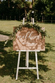 a welcome sign with greenery on it in front of a grassy field and trees