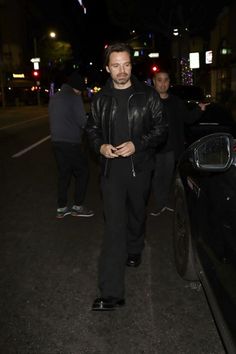 a man in black jacket and pants walking next to a car on street at night