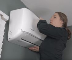 a woman in black shirt holding up a white air conditioner on top of a wall