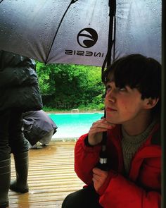 a young man sitting under an umbrella next to a swimming pool with trees in the background