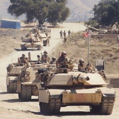 several tanks driving down a dirt road with people in the back ground and onlookers behind them