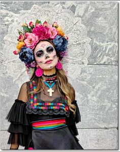 a woman with face paint and flowers in her hair wearing a black dress, standing against a wall