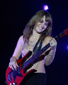 a woman holding a red guitar in her right hand and smiling while standing on stage