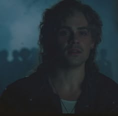 a man with long hair wearing a black leather jacket and necklace in a dark room