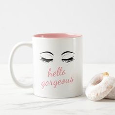 a pink and white coffee mug with eyelashes on it next to a donut that says hello gorgeous