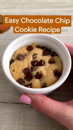 a hand holding a white bowl filled with chocolate chip cookie recipe on top of a wooden table