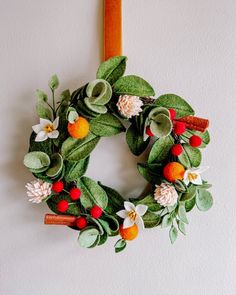 a green wreath with oranges, white and red flowers hanging on a wall next to a wooden stick