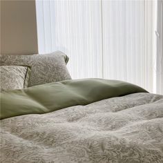 an unmade bed in front of a window with white vertical blinds and green comforter