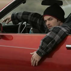 a man sitting in the driver's seat of a red car with his hand on the steering wheel