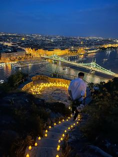 a man sitting on top of a hill with candles in front of him and the city lights behind him