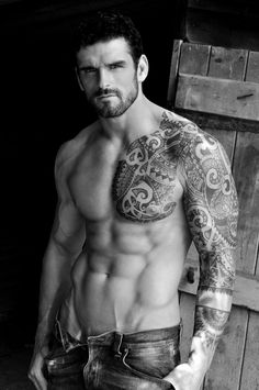 a shirtless man with tattoos standing in front of a wooden door and holding his hands on his hips