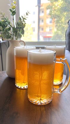 two mugs filled with beer sitting on top of a table next to a vase