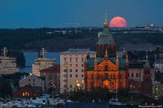 the full moon is setting over a city with tall buildings and spires in the foreground