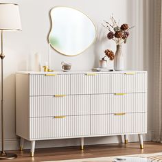 a white dresser with gold handles and drawers in front of a large round mirror on the wall