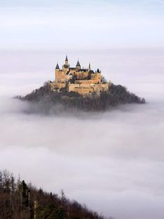 an old castle in the middle of some foggy sky with trees on top and clouds below