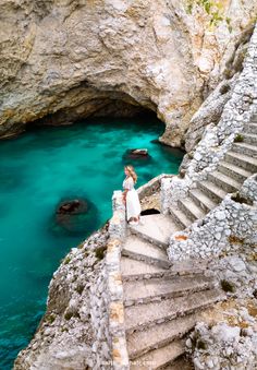 a woman is standing on some steps near the water and rocks, looking down at her