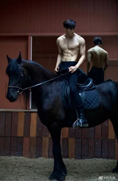 a shirtless man riding on the back of a black horse in an indoor arena