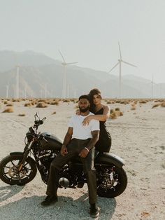 a man and woman sitting on a motorcycle in the desert with wind mills behind them