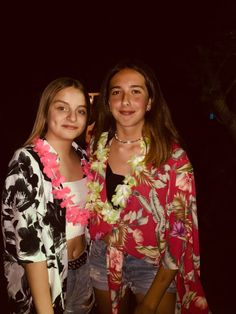 two young women standing next to each other in front of a dark background, one wearing a flower lei