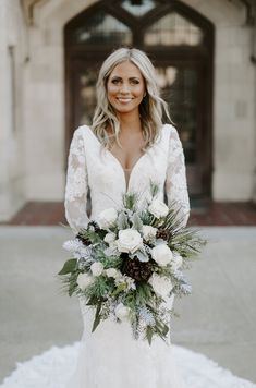 a woman standing in front of a building holding a bouquet of white flowers and greenery