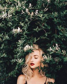 a woman with her eyes closed standing in front of some flowers and greenery looking at the camera