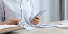 a woman sitting at a table holding a cell phone and looking at some bills in front of her