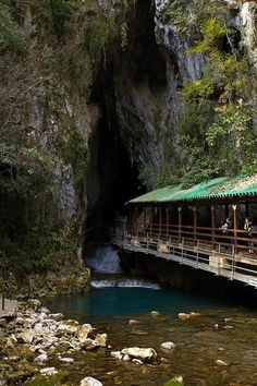 people are walking across a bridge over a river in a cave like area that is surrounded by rocks and greenery