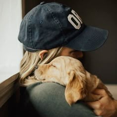 a woman holding a dog wearing a baseball cap on top of her head while sitting in front of a window