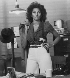 an old photo of a woman standing in front of a desk holding a camera and looking at the camera