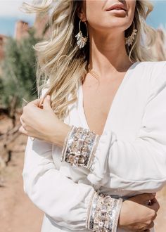 a woman with blonde hair wearing large earrings and a white dress standing in the desert