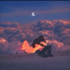 a man sitting on the ground in front of clouds with a half moon behind him