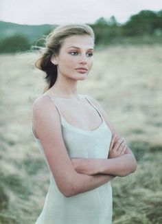 a woman standing in a field with her arms crossed and looking at the camera while wearing a white dress