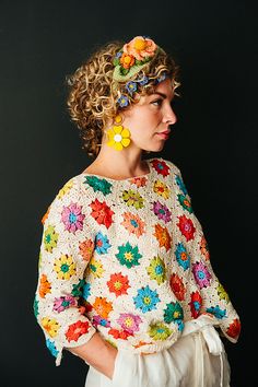 a woman with curly hair wearing a colorful top and white pants, standing in front of a black background