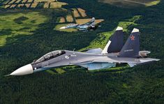 two fighter jets flying in the sky above trees and land below them are green fields