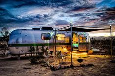 an airstream sits in the desert under a cloudy sky at dusk with chairs and tables around it